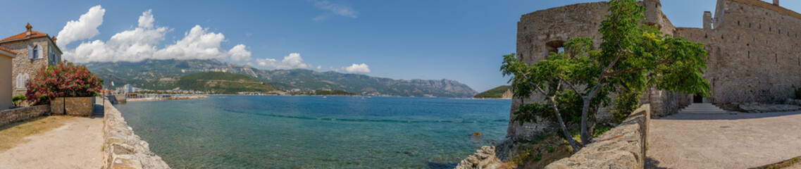 Naklejka premium Breathtaking View of a Coastal Town in Montenegro, Showcasing Crystal Clear Waters, Ancient Stone Walls, Vibrant Bougainvillea, and Majestic Mountains Under a Bright Blue Sky