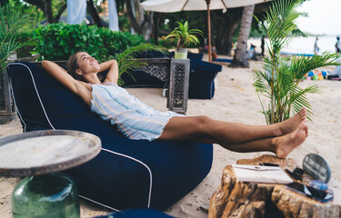Cheerful woman stretches legs on beach lounger, smiling while surrounded by tropical decor,...