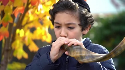Young boy wearing a kippah blows a shofar outdoors during autumn, with colorful leaves in the background.