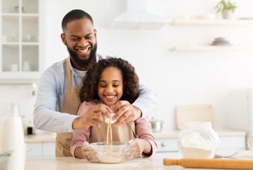 Home Bakery. Excited African American man adding egg to cookies dough, cheerful daughter watching him, learning how to cook together with dad, making pie on father's day, copy space