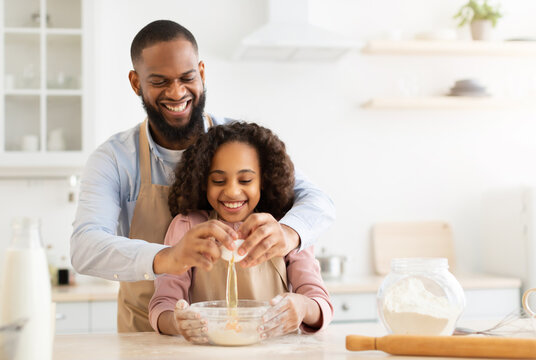 Home Bakery. Excited African American man adding egg to cookies dough, cheerful daughter watching him, learning how to cook together with dad, making pie on father's day, copy space
