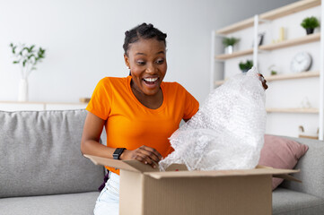 Happy black woman unboxing cardboard parcel, emotionally reacting to successful shopping, taking...