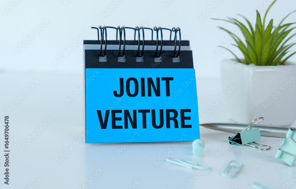 Poster a blue notebook with the word joint venture written on it. the notebook is on a table with a pen and