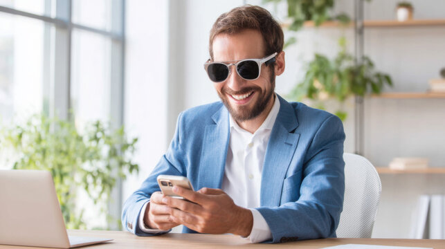 A smiling man in sunglasses sits at a desk, looking at his phone in a bright office filled with plants.