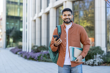 Obraz na płótnie Canvas Cheerful young adult with backpack, books, and smartphone, standing outdoors near a modern building. The scene conveys education, connection, and a vibrant energy of student life.