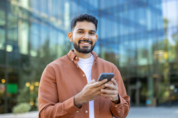 A cheerful man standing outdoors uses his smartphone in a professional urban setting. His happiness...