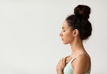 Profile portrait of calm young woman with hands on chest and closed eyes, side view shot of peaceful millennial female meditating or praying while standing over white background indoors, copy space