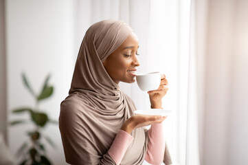 Coffee time. Gorgeous black woman in hijab sipping hot aromatic drink near window at home. Lovely...