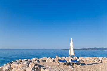 Minimalist summer scene: Two empty beach chairs and an umbrella on a rocky coast with a calm blue...