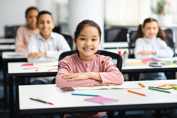 Elementary Education. Portrait of happy small asian girl sitting at table in classroom at primary school with group of classmates and posing, looking at camera and smiling. Studying And Learning