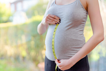 Woman measuring her pregnant belly with a tape measure in a sunny garden setting during the day