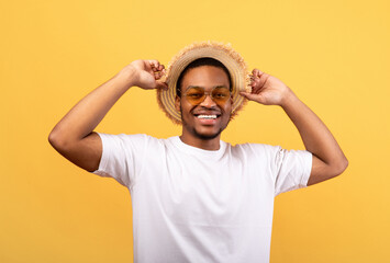 Portrait of handsome African American man in casual clothes, straw hat and sunglasses on yelow studio background. Millennial black guy wearing summer outfit, ready for beach vacation