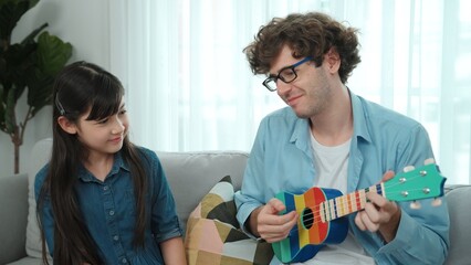 Caucasian attractive dad playing ukulele while young smart daughter listening while move to song. Skilled father play guitar while listen music and dancing at sofa. Family activity concept. Pedagogy.