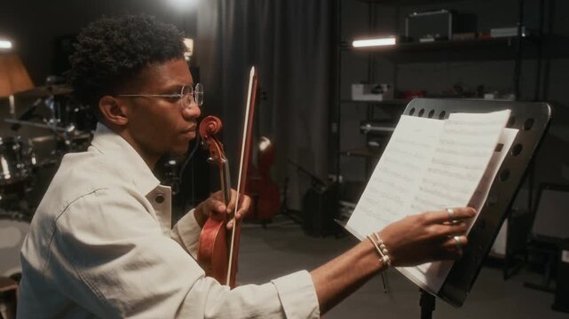 Side view shot of young African American man as modern violinist reading sheet music and turning pages while practicing in studio with cinematic lighting, copy space