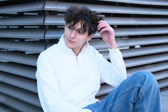 Casual teen guy in blue jeanse sitting on stone fence against urban street