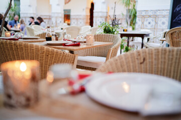 Cozy outdoor restaurant setting with wicker chairs and wooden tables, featuring candles and tableware, creating a warm and inviting atmosphere.