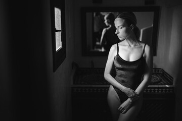 A woman in a dark swimsuit stands in a dimly lit room, gazing out a window. The room features a...