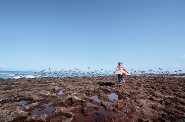 A young woman chases a flock of birds on a rocky seaside under a sunny blue sky, evoking feelings...
