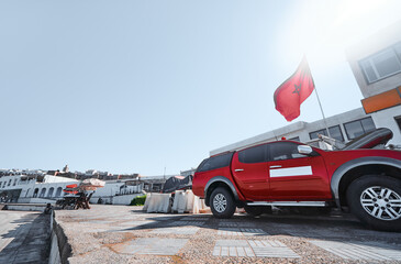 A vibrant red pickup truck is parked near white buildings and a waving Moroccan flag under clear...
