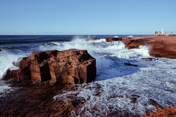 Dramatic scenery of waves crashing onto rugged cliffs under a clear blue sky. A lighthouse stands...