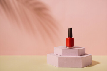 A bottle of red nail polish with a black cap, displayed on a modern pink hexagonal podium. This...
