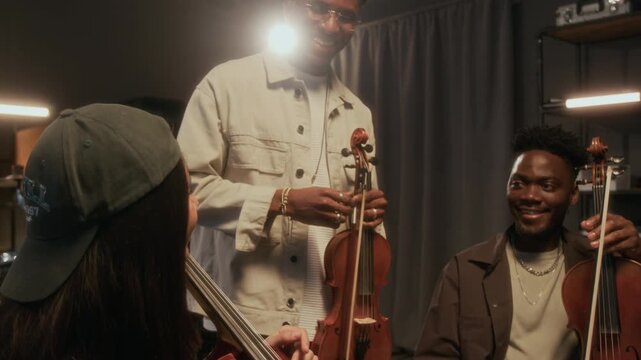 Group of young people as string trio enjoying conversation during music rehearsal together and smiling cheerfully, backlit by studio lights