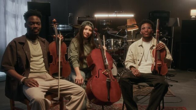 Front view shot of young people as string trio posing with musical instruments violin and cello while looking at camera in recording studio