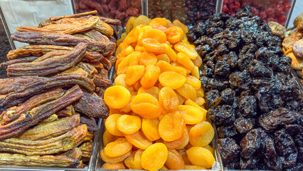 A close-up view of a variety of stacked dried fruits, including banana slices, orange apricots, and black prunes. The distinct textures and colors of the fruits are evident in the display.