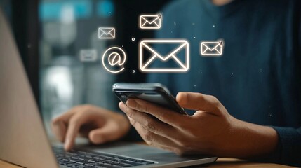 Man using smartphone while working on laptop in modern office setting with email notifications appearing on screen
