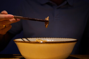 A hand with chopsticks lifts a tender piece of meat above a ceramic bowl of noodles, capturing the quiet anticipation and intimate ritual of enjoying a warm meal.