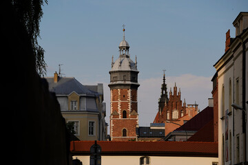 Tall brick tower with silver roof rises among rooftops and gothic spires, framed by evening light,...