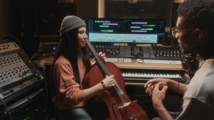Side view shot of young woman playing cello in recording studio with music producer listening during production of new instrumental album