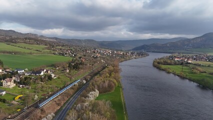 Drone view of a winding river with a passing train, green hills, and small villages. Scenic countryside landscape with mountains in the background under dramatic cloudy sky.