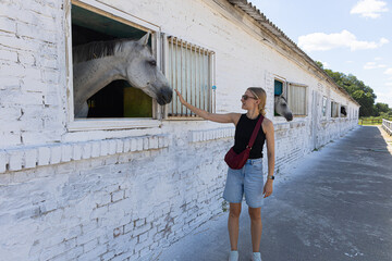 Woman petting white horse in stable yard on sunny day