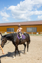Young child in protective helmet practicing horseback riding on training horse under guidance of...