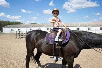 Smiling little girl riding a black horse at equestrian training ground