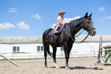 Smiling little girl riding a black horse at equestrian training ground