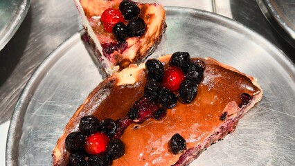 Two slices of homemade cheesecake with blueberries and cherries on a metal plate — an appetizing dessert for advertising baked goods, menus, culinary blogs, and seasonal content.