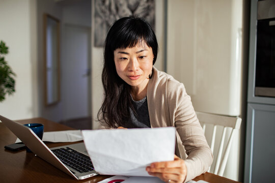 Woman reviewing utility bill at home while using laptop in bright kitchen