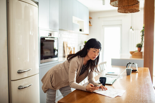 Woman reviewing utility bill at home while using laptop in bright kitchen