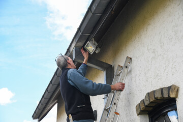 Electrician checks the settings of a motion sensor LED floodlight while standing on a ladder, the...