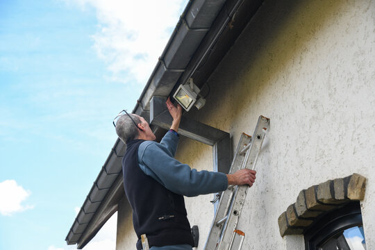 Electrician checks the settings of a motion sensor LED floodlight while standing on a ladder, the light is used when needed, saving energy during an energy crisis
