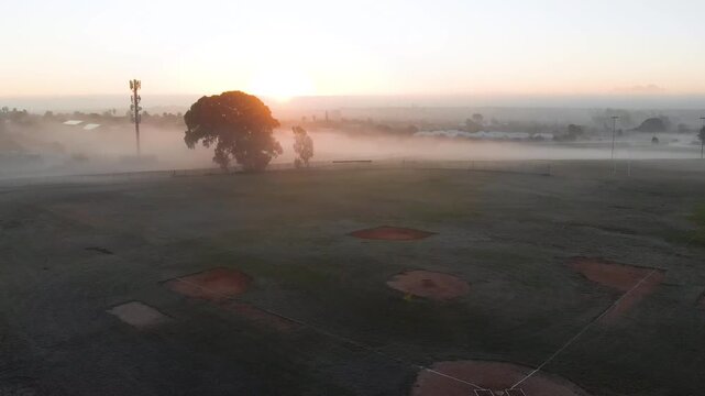 Sun is rising above horizon, lifting fog, sending golden light and revealing baseball infields