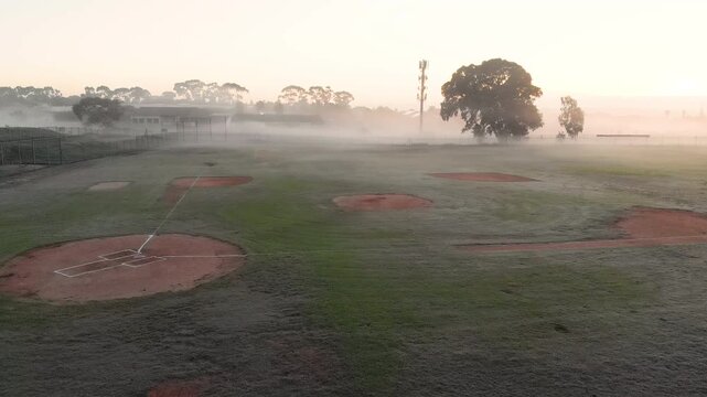 Camera tilting downward from fence to reveal baseball infields as mist lifts under sunrise glow