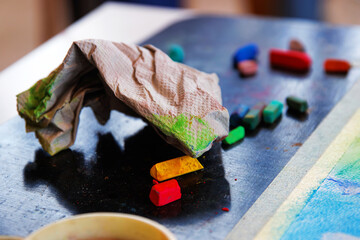 Crumpled paper towel with colorful pastel chalk stains placed on a dark art table, surrounded by scattered chalk sticks in vibrant colors
