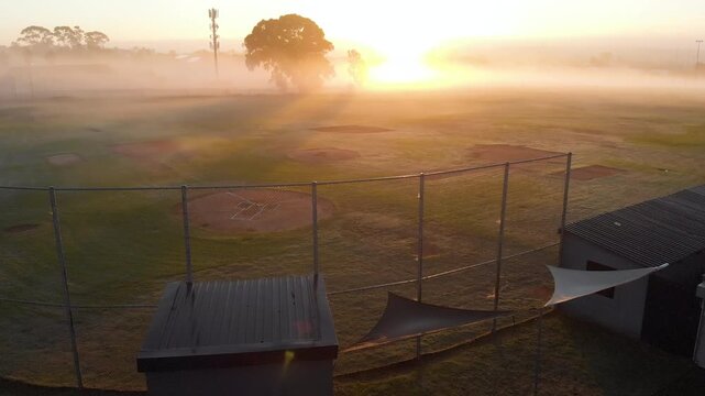 Rising sun is driving camera hovering behind chainlink fence tilting down showing baseball infields