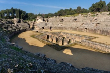 Ancient Roman Amphitheater itálica: A grand stone amphitheater stands proudly against a backdrop of verdant trees, the arena's walls weathered by time and history.