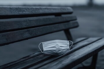 A disposable face mask rests forlornly on a wet, dark wooden park bench in a somber, overcast setting.