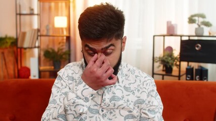 Indian young man sitting at home, removing glasses and rubbing sore eyes, feeling strained and tired from remote work. Arabian guy experiencing discomfort while staying on sofa in modern apartment. - Powered by Adobe