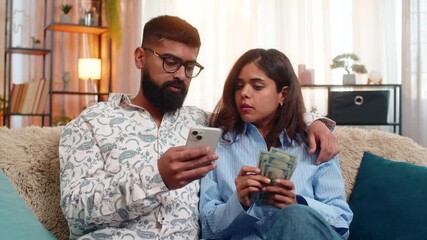 Indian couple man and woman sitting on home sofa holding money dollar cash and smartphone while planning purchases and family budget. Discussing future expenses, savings and income from salary indoors - Powered by Adobe